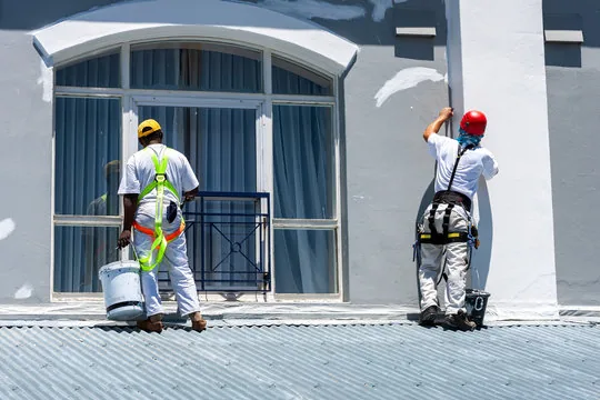 Two harnessed painters working on a commercial building facade from a metal roof